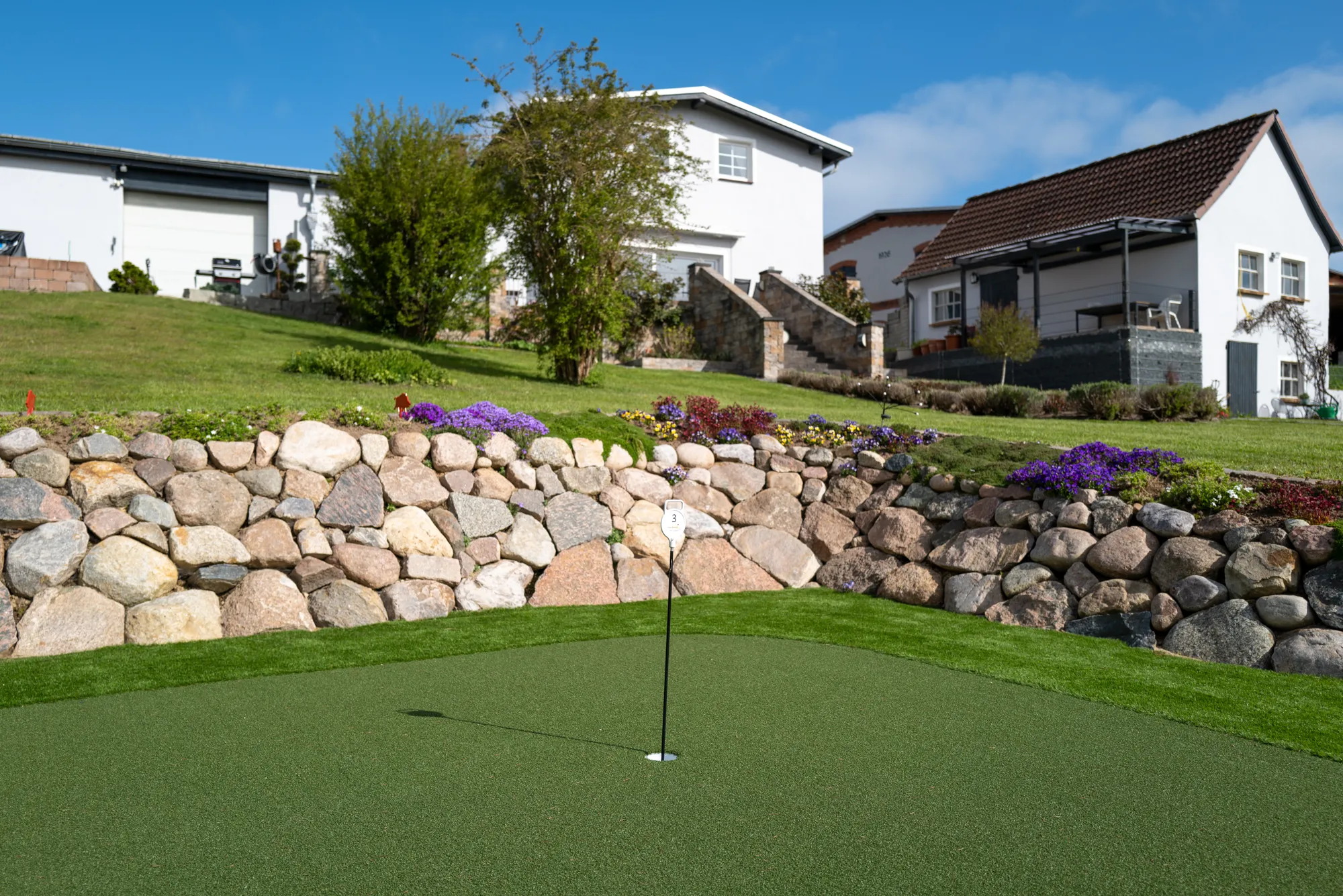 Putting-Green mit Fahne, eingefasst von einer Natursteinmauer, im Garten eines Wohnhauses
