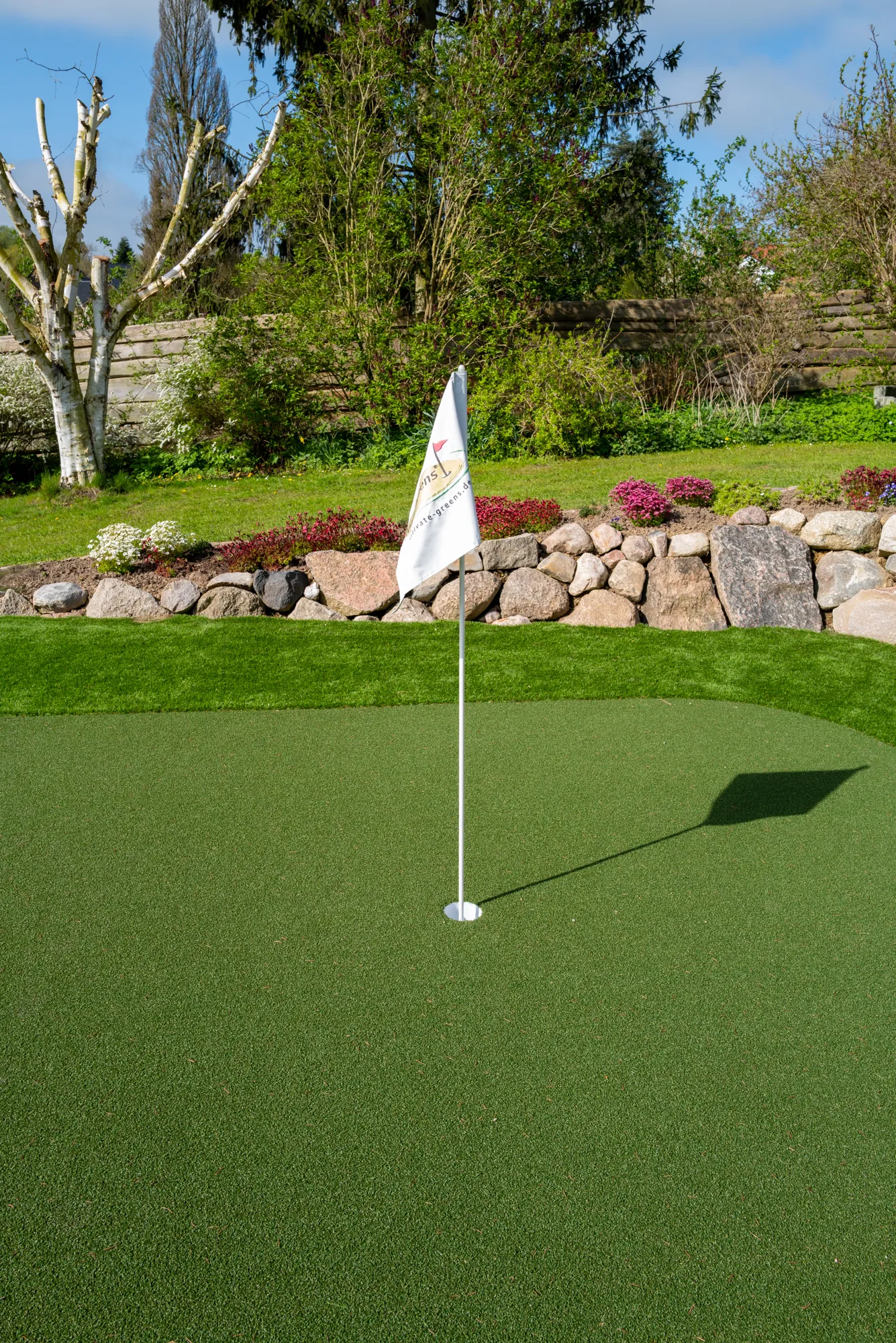 Golfgreen mit Private-Greens-Fahne vor Natursteinmauer und Gartenbepflanzung