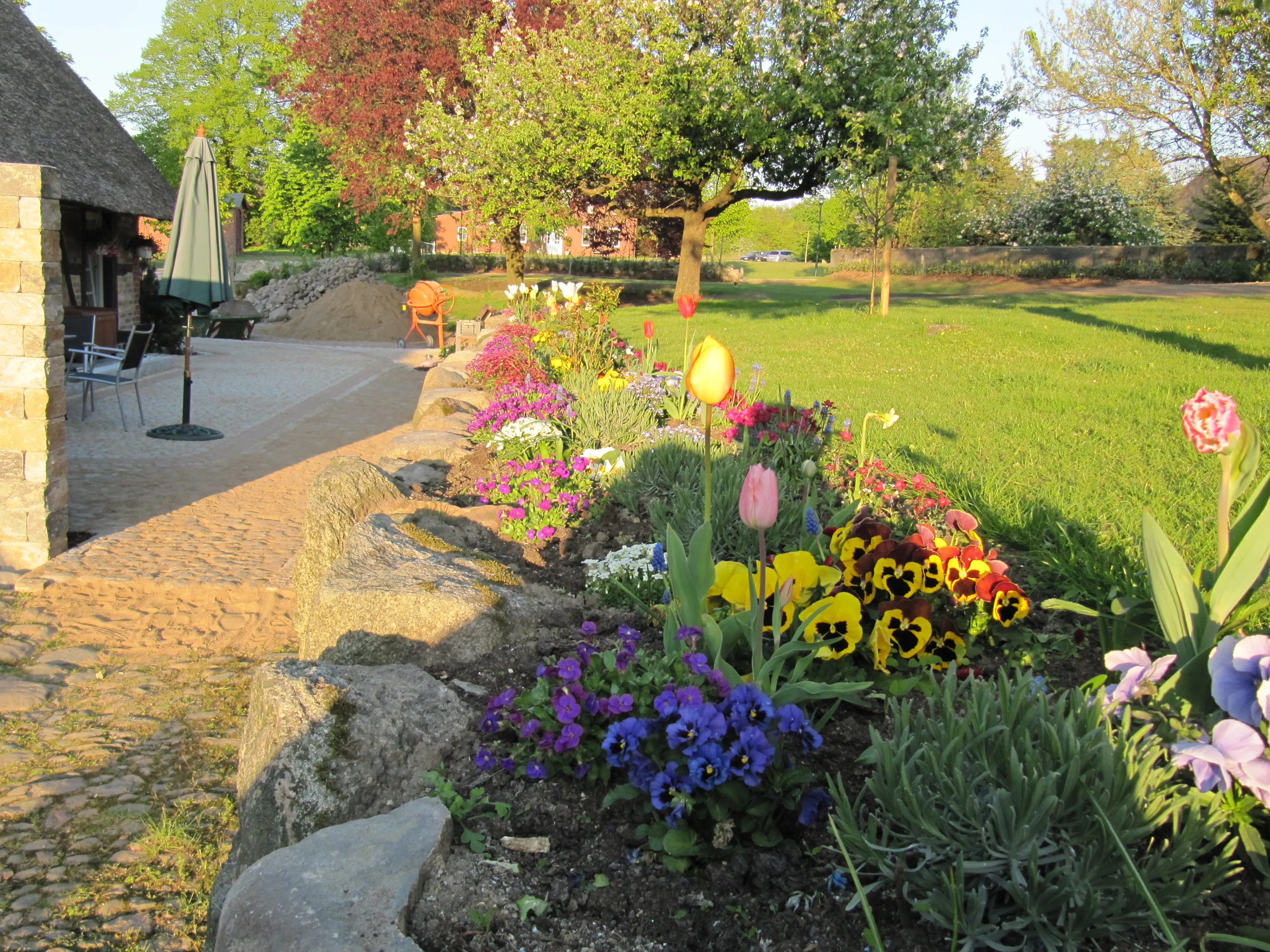 Farbenfrohes Frühlingsbeet mit Tulpen, Stiefmütterchen und Stauden, eingefasst von Natursteinen neben einer Terrasse